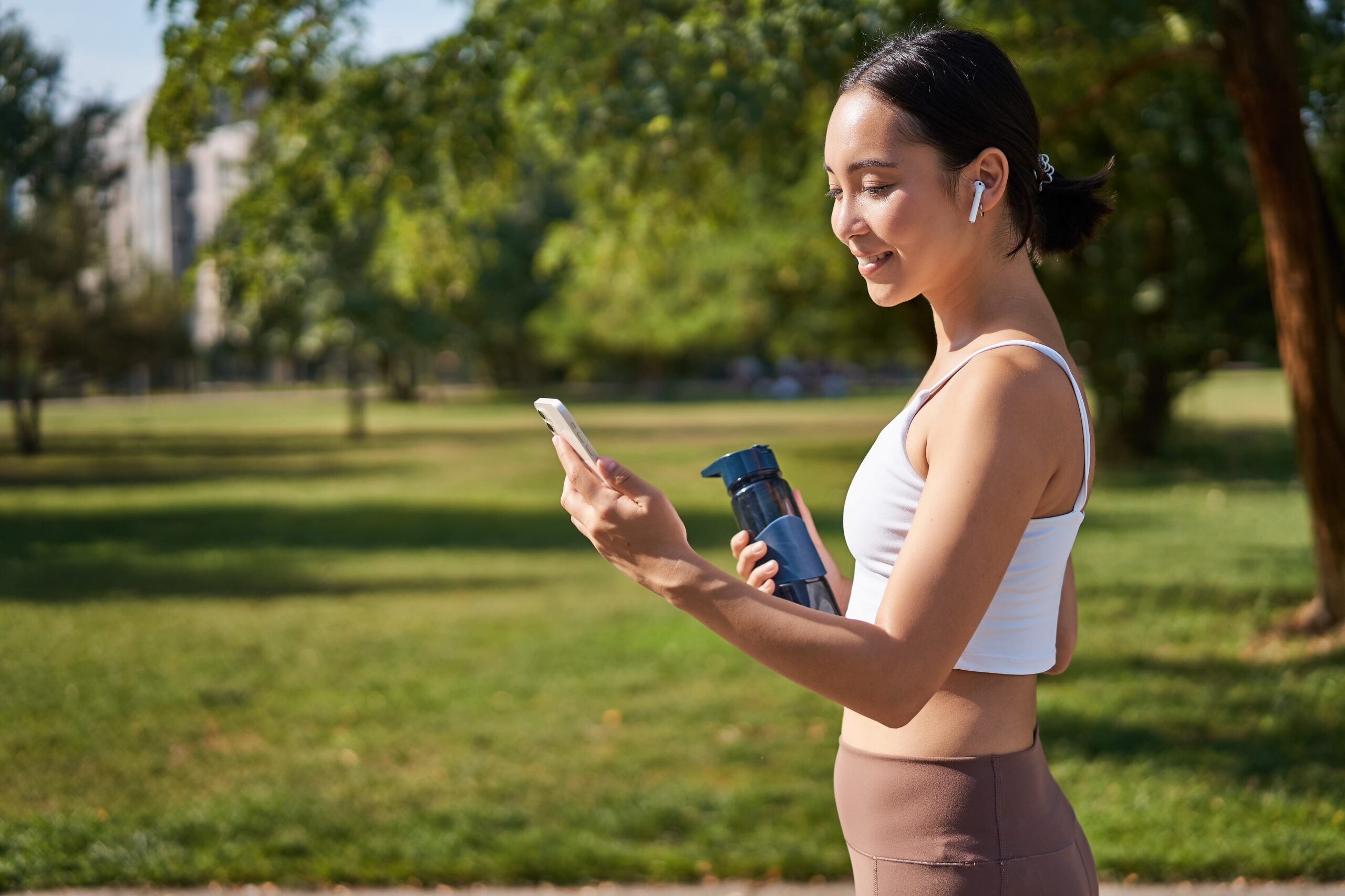 fitness girl runner drinking water looking at smartphone in the park 