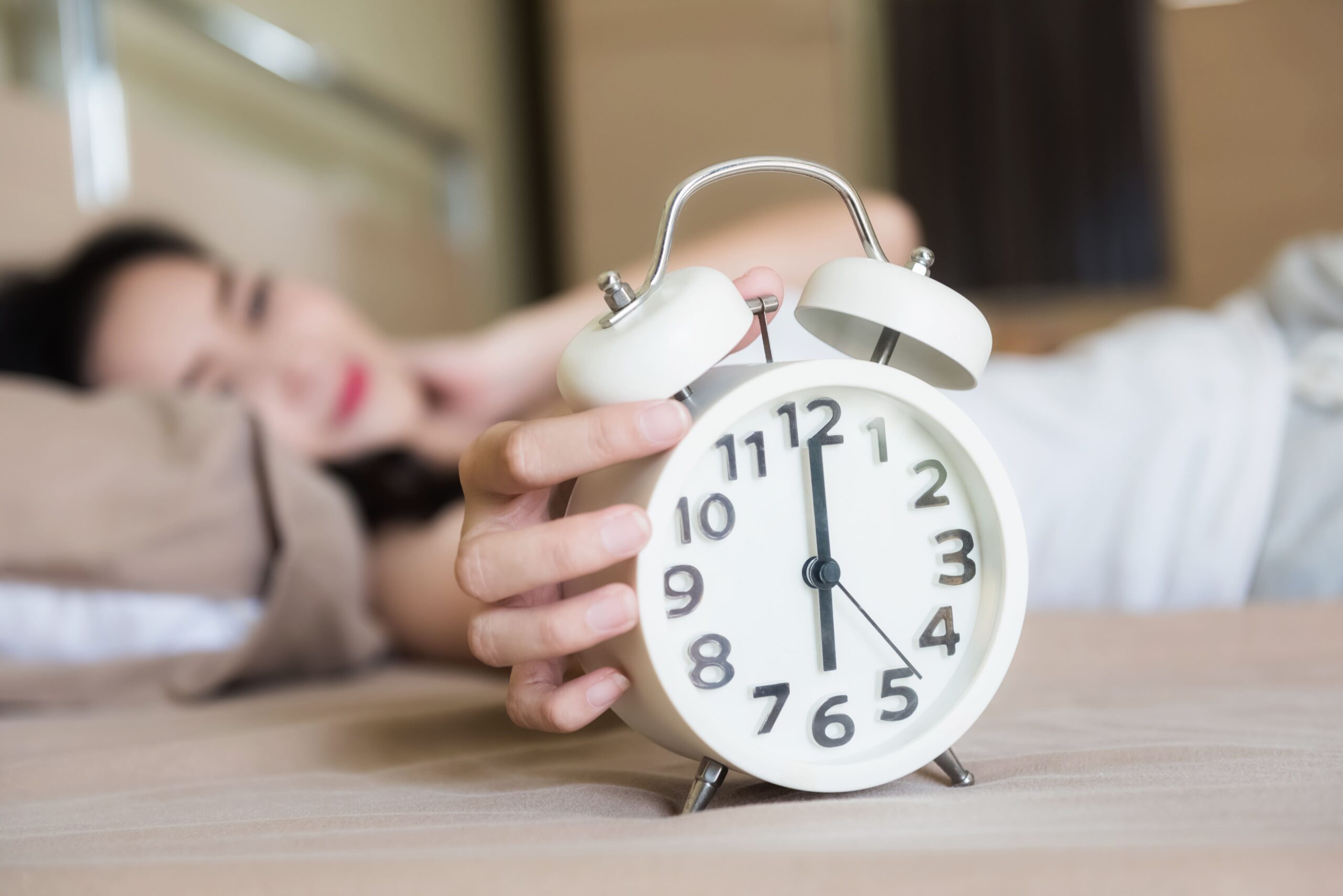 Person lying in bed reaching to turn off a white alarm clock