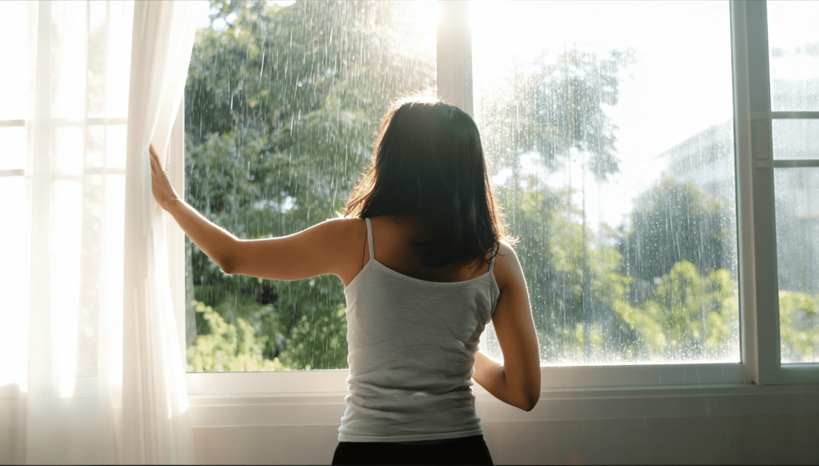 Woman opening curtains to let in natural light on a rainy morning, a simple way to support mental health during the rainy season.