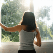 Woman opening curtains to let in natural light on a rainy morning, a simple way to support mental health during the rainy season.