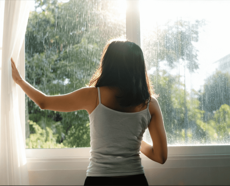 Woman opening curtains to let in natural light on a rainy morning, a simple way to support mental health during the rainy season.