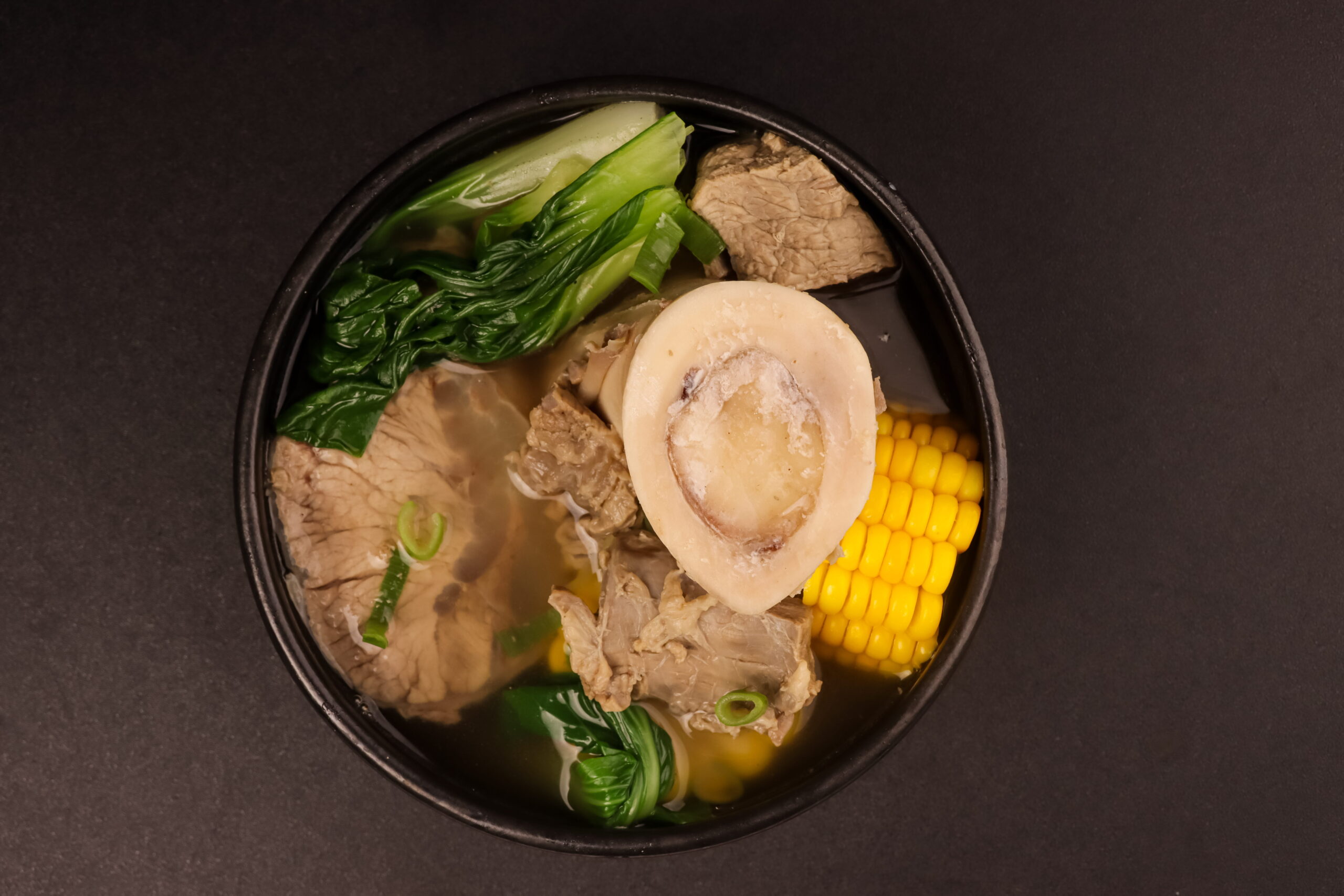 A steaming bowl of Filipino beef bulalo soup with tender beef shank, corn on the cob, and leafy greens.