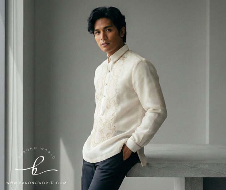 Man in cream jusi barong with intricate embroidery, dark trousers, leaning against a stone ledge.
