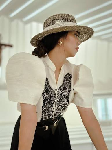 A woman wearing an embellished straw hat and an off-white modern Filipiniana top with black embroidery and puffed sleeves.