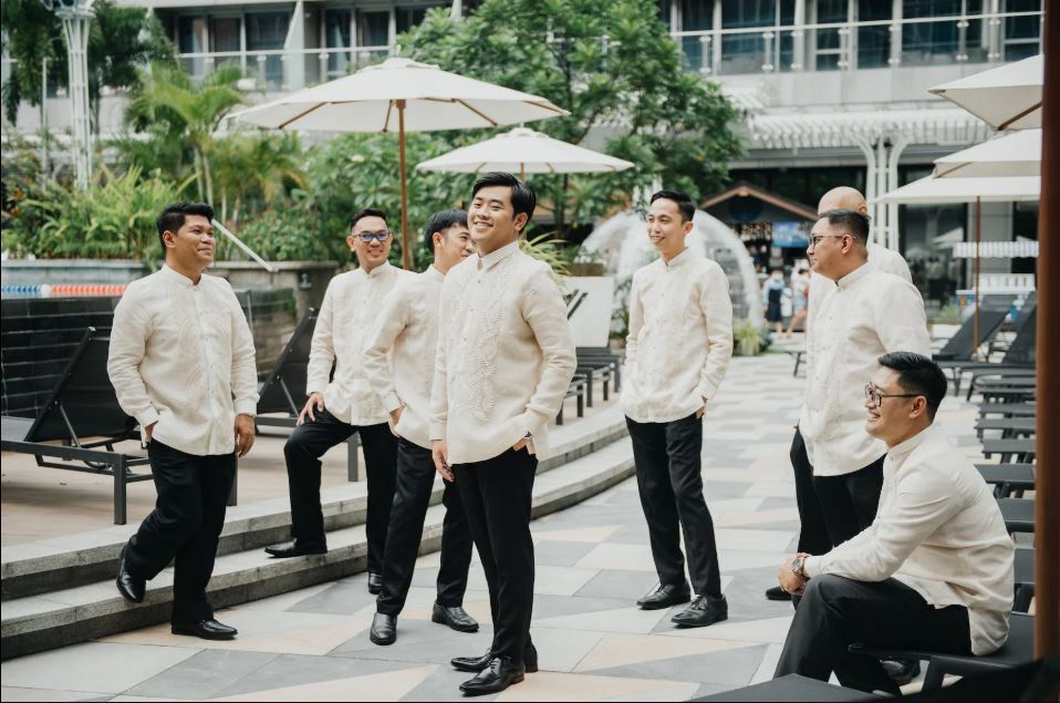 A group of groomsmen wearing traditional embroidered beige Barong Tagalog outfits outdoors near a pool area.