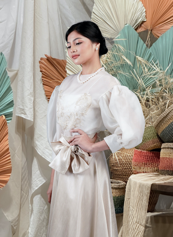 Woman in ivory embroidered Filipiniana dress with puff sleeves and a bow sash, pearl necklace, styled portrait.