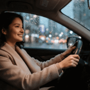 A smiling woman in a beige coat drives a car at night as rain falls on the window, with blurry city lights outside.