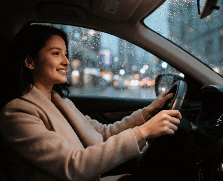 A smiling woman in a beige coat drives a car at night as rain falls on the window, with blurry city lights outside.