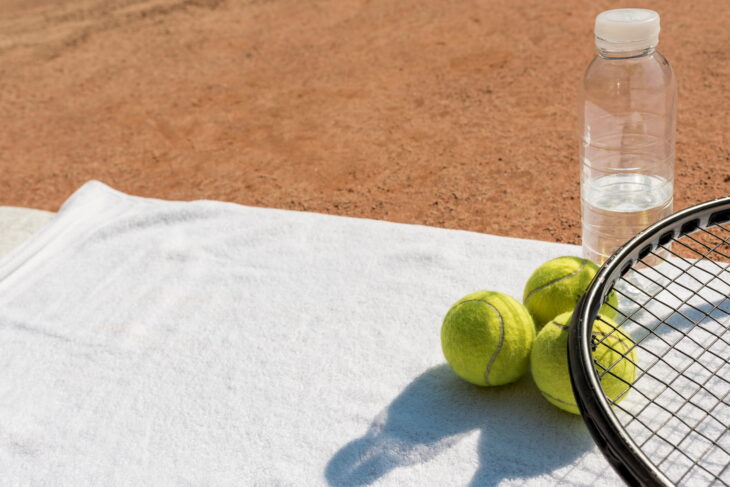 Tennis racket, three tennis balls, and a water bottle on a white towel in an outdoor tennis court